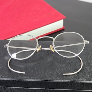 May include: Silver-toned metal eyeglasses featuring round lenses and thin wire frames. The eyewear includes curved cable temples designed to hook behind the ears and small, gold-colored nose pads attached to the bridge for a secure fit.
