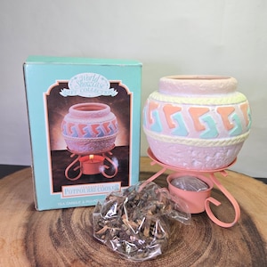 May include: A light pink ceramic potpourri cooker with a textured base and a geometric pattern in peach and light blue, resting on a pink metal stand. The set includes a box labeled World Showcase Gift Collection and a bag of dried potpourri.
