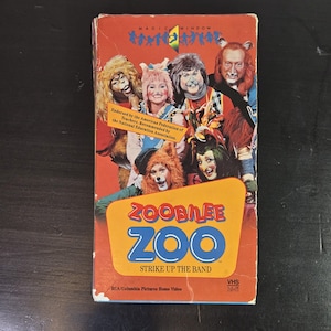 Puede incluir: Una cinta VHS de Zoobilee Zoo Strike Up The Band con una portada roja que muestra a un grupo de personajes disfrazados con trajes inspirados en animales. El título aparece en letras grandes azules y rojas, con texto sobre avales educativos.