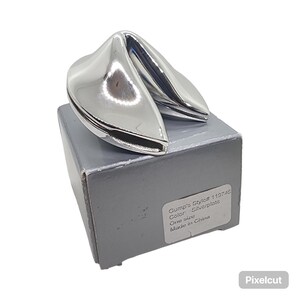 May include: A silver-plated fortune cookie sculpture with a polished, reflective surface resting on a grey cardboard box. The box features a white label with the text: Gump's Style# 119746, Color: Silverplate, One size, Made in China.