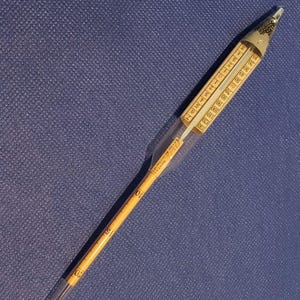 May include: A vintage hydrometer with a clear glass body and a yellow paper scale. The scale is marked with numbers and lines. The hydrometer is designed to measure the specific gravity of liquids.