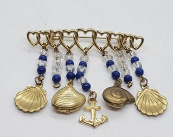 May include: Gold-toned brooch featuring a row of heart-shaped links. Dangling from the hearts are strands of clear and blue beads, and charms shaped like seashells, a nautilus shell, and an anchor. A nautical-themed accessory.
