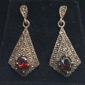 May include: A pair of silver dangle earrings with a red gemstone in the center of each earring. The earrings are decorated with a geometric pattern of small silver beads.