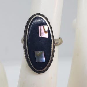 May include: Silver ring with an oval black stone inlay, accented with two iridescent squares. The ring has a twisted rope-like border and a simple band. The ring is displayed on a white background.