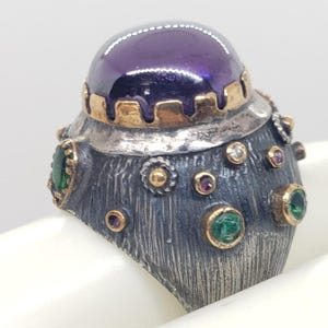 May include: A unique ring featuring a large, domed purple gemstone. The ring has a silver band with gold accents and is adorned with smaller green and red gemstones. The design is intricate and detailed, showcasing a blend of textures and colors.