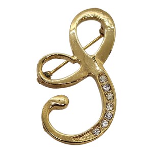 May include: Gold-toned metal brooch in the shape of the letter "S". The brooch is embellished with clear, sparkling rhinestones along the curve of the letter. The pin closure is visible on the back. A stylish accessory for jackets, scarves, or bags.