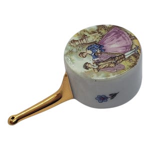 May include: A small, white ceramic pot with a gold handle. The top of the pot features a painted scene of two people in period clothing in a garden. A small blue flower is painted on the side.