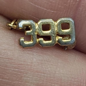 May include: A small, gold-toned metal charm with the number "399" in a blocky font. The charm has a small loop at the top for attachment and shows signs of wear. The background is a blurred view of a skin tone.