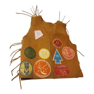 May include: A tan leather vest adorned with various colorful patches. The patches feature text and graphics related to camping, awards, and events. The vest has fringe along the sides and is likely vintage.