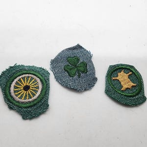 May include: Three embroidered patches on denim fabric. One features a yellow and white wheel design, another a green shamrock, and the third a tan animal within a green circle. The patches have a frayed edge.