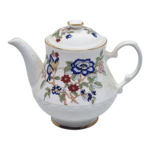 May include: White ceramic teapot with a lid, featuring a floral and butterfly design in blue, red, and pink. The teapot has a gold trim around the base and lid. The handle and spout are white.