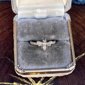 May include: A silver-toned ring featuring a detailed bee ornament centered on the band. The bee has textured wings and a segmented body. The ring is displayed inside a small, rectangular jewelry box lined with grey velvet fabric.