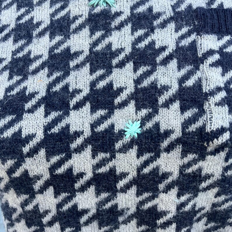 Puede incluir: Primer plano de un su&eacute;ter de punto con un cl&aacute;sico estampado de pata de gallo en azul marino y blanco roto. El su&eacute;ter presenta peque&ntilde;os detalles florales bordados en azul claro. La tela parece suave y texturizada, con puntadas de punto visibles.