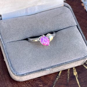 May include: A silver ring with a pear-shaped, pink gemstone. The ring is set in a gray velvet ring box. The gemstone is a vibrant pink color and is the focal point of the ring.