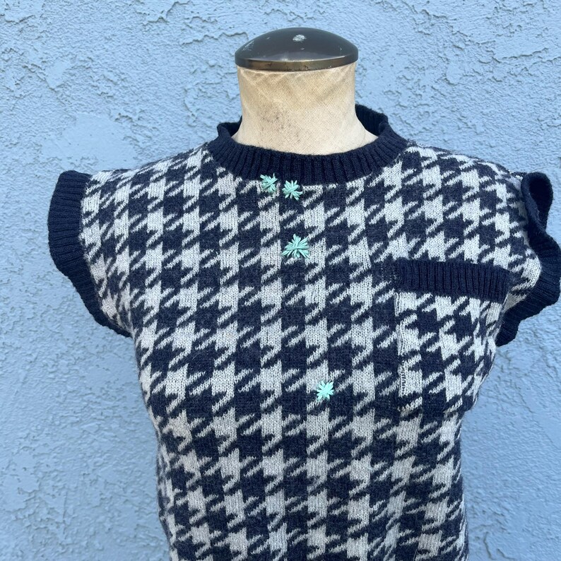 Puede incluir: Un su&eacute;ter sin mangas con estampado de pata de gallo en azul marino y crema. El su&eacute;ter tiene cuello redondo, un peque&ntilde;o bolsillo y tres botones en forma de flor verde claro. Los bordes est&aacute;n ribeteados en azul marino.