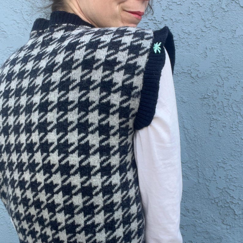 Puede incluir: Un chaleco con estampado de pata de gallo en blanco y negro con ribete azul marino. El chaleco tiene un cuello alto y un peque&ntilde;o dise&ntilde;o bordado en verde claro en el hombro. El chaleco se lleva sobre una camisa blanca de manga larga.