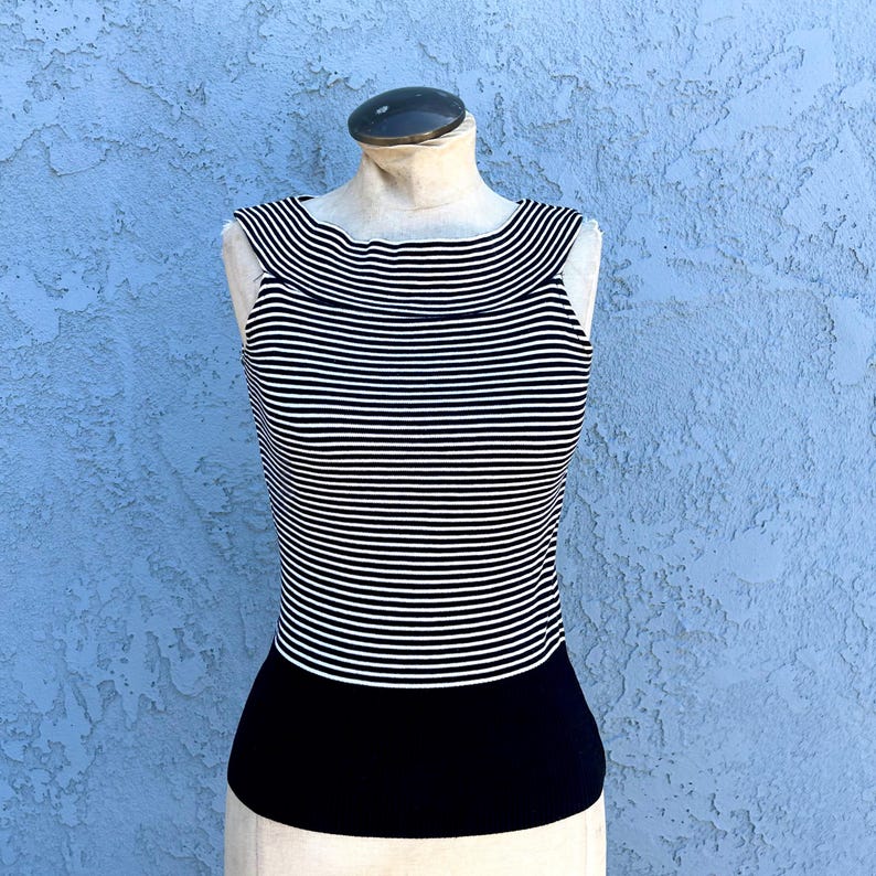 May include: A sleeveless top with horizontal black and white stripes on the upper portion and a solid black lower section. The top is displayed on a mannequin against a textured blue background.