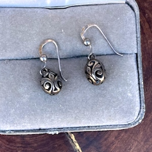 May include: A pair of silver-toned drop earrings. Each earring features a small, oval-shaped pendant with an intricate, openwork design. The earrings are displayed in a gray jewelry box, showcasing their delicate craftsmanship and vintage style.