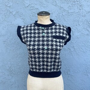 Puede incluir: Un su&eacute;ter sin mangas de pata de gallo en azul marino y gris claro, exhibido en un maniqu&iacute;. El su&eacute;ter presenta un peque&ntilde;o bolsillo, botones decorativos y mangas con volantes. El cuello y el dobladillo son de color azul marino.