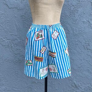 May include: Elastic-waist shorts featuring a pattern of vertical white and bright blue stripes. The fabric is decorated with various rectangular patches and graphic illustrations, including text such as Venice, California, and other small colorful designs.