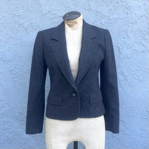 May include: A charcoal gray blazer featuring a single-button closure, notched lapels, and two front flap pockets. The garment is displayed on a mannequin, showcasing long sleeves and a structured fit with a textured, wool-like fabric appearance.