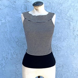 May include: A sleeveless top with horizontal black and white stripes. The top has a scoop neckline and a solid black ribbed bottom. The garment is displayed on a mannequin against a textured blue background.