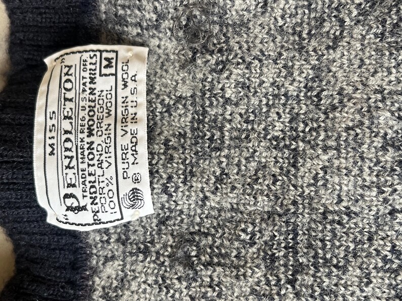 Puede incluir: Primer plano de un su&eacute;ter de punto gris y blanco con una etiqueta que dice "Pendleton" y "100% lana virgen". La etiqueta tambi&eacute;n indica que el su&eacute;ter es talla M y est&aacute; hecho en los EE. UU.