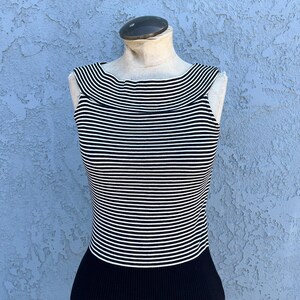 May include: A sleeveless top with horizontal black and white stripes. The top has a wide neckline and a black ribbed bottom. The garment is displayed on a mannequin against a textured blue background.
