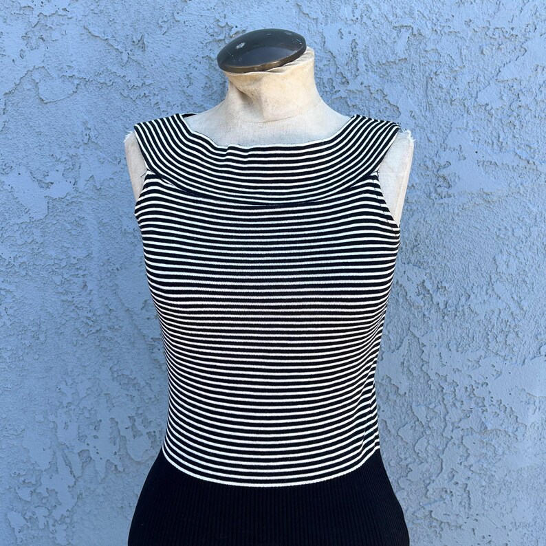 May include: A sleeveless top with horizontal black and white stripes. The top has a wide neckline and a black ribbed bottom. The garment is displayed on a mannequin against a textured blue background.