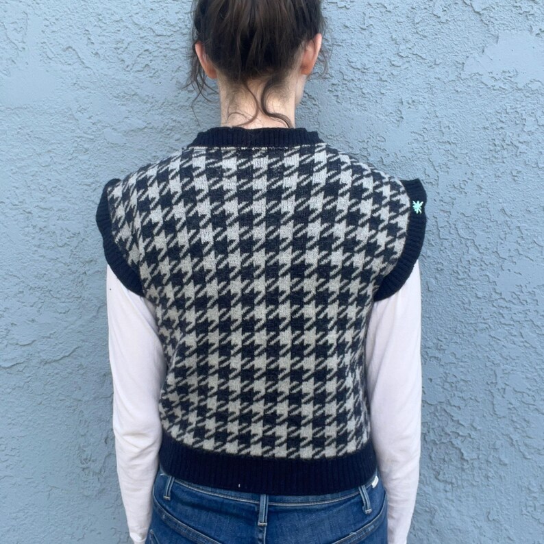 Puede incluir: Un chaleco sin mangas con estampado de pata de gallo en blanco y negro, con ribete azul marino. El chaleco se lleva sobre una camisa blanca de manga larga y vaqueros azules. Un peque&ntilde;o detalle bordado verde es visible en el hombro.