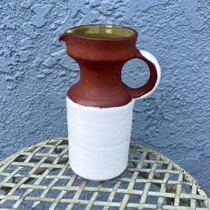 May include: A handmade ceramic pitcher with a white base, a reddish-brown midsection, and a green interior. The pitcher has a handle and a spout. The pitcher is sitting on a metal table with a lattice design.