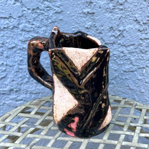 May include: A handmade ceramic mug with a unique, textured design. The mug features a black, brown, and pink color scheme with a geometric pattern. The handle is integrated into the design, and the mug sits on a metal surface.