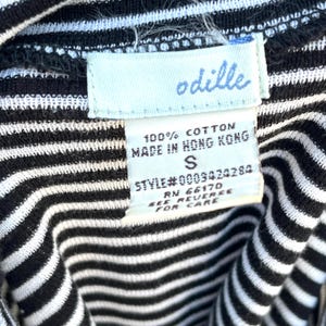 May include: Close-up of a black and white striped garment with a tag. The tag reads "odille" in light blue script. The tag also states "100% Cotton, Made in Hong Kong, S, Style#0009424284, RN 66170, See Reverse For Care."