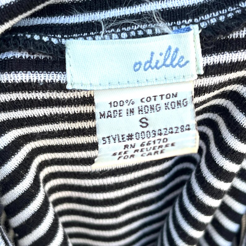 May include: Close-up of a black and white striped garment with a tag. The tag reads "odille" in light blue script. The tag also states "100% Cotton, Made in Hong Kong, S, Style#0009424284, RN 66170, See Reverse For Care."
