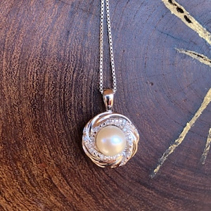 May include: A silver pendant necklace featuring a round, creamy-white pearl at the center, surrounded by a swirling, textured silver design. Small, clear stones are set around the pearl. The necklace hangs on a delicate silver chain.