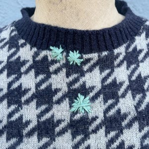 Puede incluir: Primer plano de un su&eacute;ter con estampado de pata de gallo en azul marino y blanco roto. El su&eacute;ter tiene un cuello de canal&eacute; azul marino y tres peque&ntilde;os detalles florales bordados en verde claro.