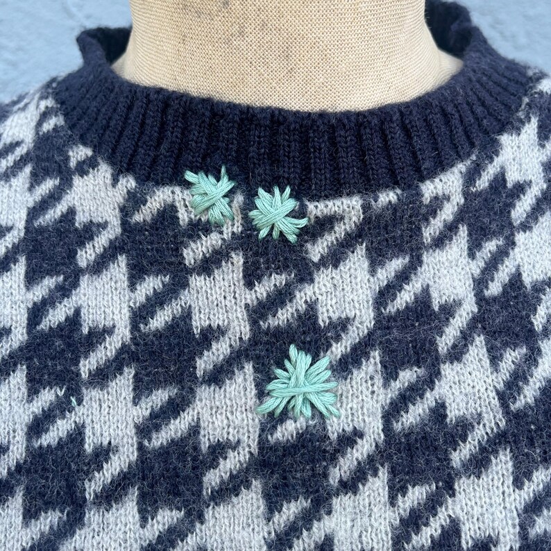 Puede incluir: Primer plano de un su&eacute;ter con estampado de pata de gallo en azul marino y blanco roto. El su&eacute;ter tiene un cuello de canal&eacute; azul marino y tres peque&ntilde;os detalles florales bordados en verde claro.