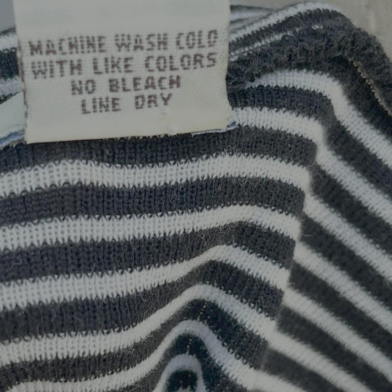May include: Close-up of a fabric tag with care instructions: "MACHINE WASH COLD WITH LIKE COLORS NO BLEACH LINE DRY." The fabric is a knit with alternating white and dark gray horizontal stripes.