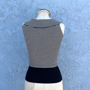 May include: A sleeveless top with horizontal black and white stripes on the upper portion and a solid black band at the waist. The top is displayed on a mannequin against a textured blue background.