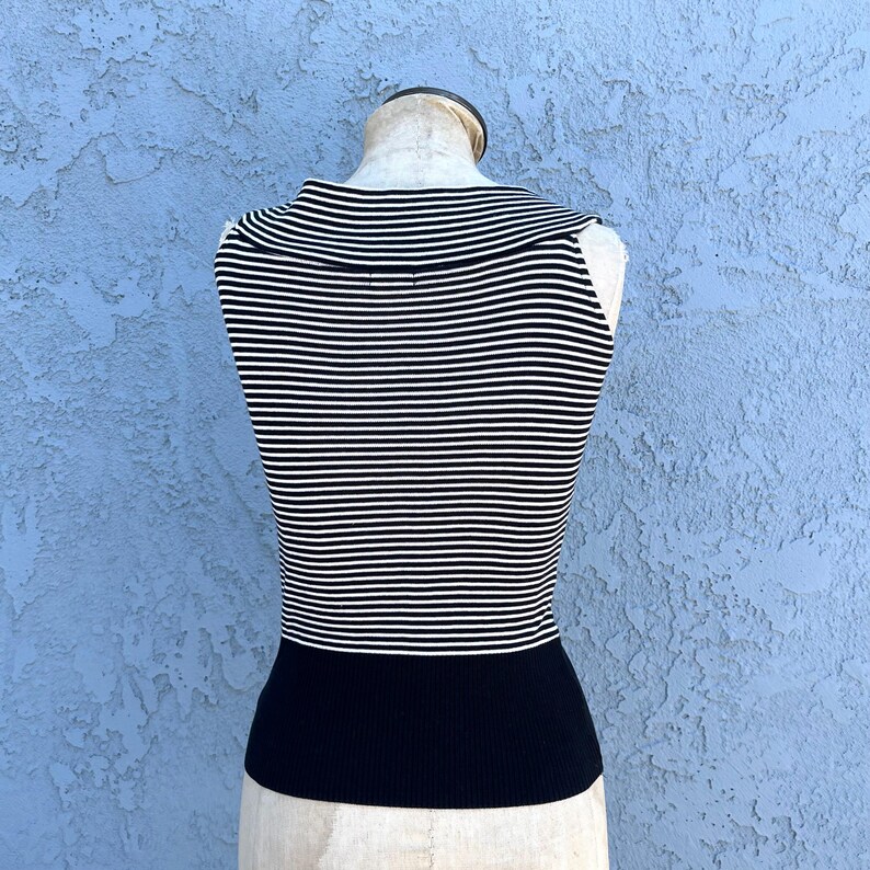 May include: A sleeveless top with horizontal black and white stripes on the upper portion and a solid black band at the waist. The top is displayed on a mannequin against a textured blue background.