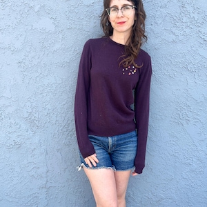 May include: A dark purple sweater with a crew neck and long sleeves. The sweater has a small, colorful embroidered design on the upper left chest. Worn with denim shorts.