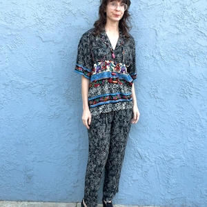 Könnte beinhalten: Ein zweiteiliges Outfit mit einem kurzärmeligen Hemd mit Knöpfen und einer passenden Hose. Das Hemd und die Hose sind schwarz mit einem weißen Blumenmuster. Das Hemd hat eine farbenfrohe bestickte Blende über der Brust und blaue Verzierungen.