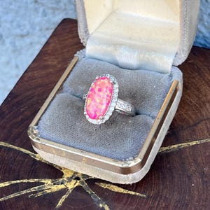 Puede incluir: Un anillo de ópalo rosa con un halo de pequeñas piedras transparentes. La gema de forma ovalada está engastada en una banda plateada y se presenta en un joyero de estilo vintage con interior de terciopelo gris.