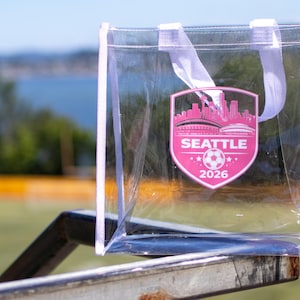 May include: A clear plastic tote bag with white trim and two white handles. The front features a pink shield-shaped graphic with a Seattle skyline silhouette, a soccer ball icon, and the text SEATTLE 2026 printed in white capital letters.
