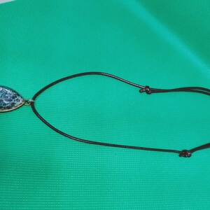 May include: A teardrop pendant necklace with a gold-coloured bezel. The pendant has a blue and white speckled design. The necklace has a black cord with adjustable knots. The background is a solid green colour.