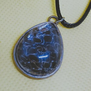 May include: A teardrop pendant with a dark blue, patterned centre, set in a silver-coloured metal frame. It hangs from a black cord necklace, a simple yet elegant accessory. The background is a light yellow.