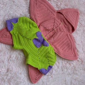 May include: Handmade crochet baby outfit in lime green with purple accents, and a matching pink crochet blanket. The outfit features a unique design, and the blanket is shaped like a star. The items are displayed on a fluffy, light pink surface.