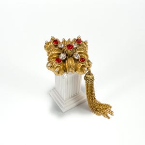 May include: A gold-toned brooch featuring a square, ornate metal design with embedded red and clear crystals, small white pearls, and a long, dangling gold chain tassel attached to one side, showcasing intricate scrollwork and textured metal details.