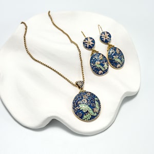 May include: A jewelry set featuring a necklace and earrings. The necklace has a round pendant with a blue floral design, and the earrings have an oval shape. All pieces have gold-colored metal and are set on a white surface.