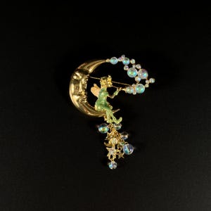 May include: A gold-tone brooch featuring a crescent moon with a face, a green fairy playing a flute, and dangling iridescent beads and stars. The fairy is seated on the moon, with a trail of bubbles extending from the flute.